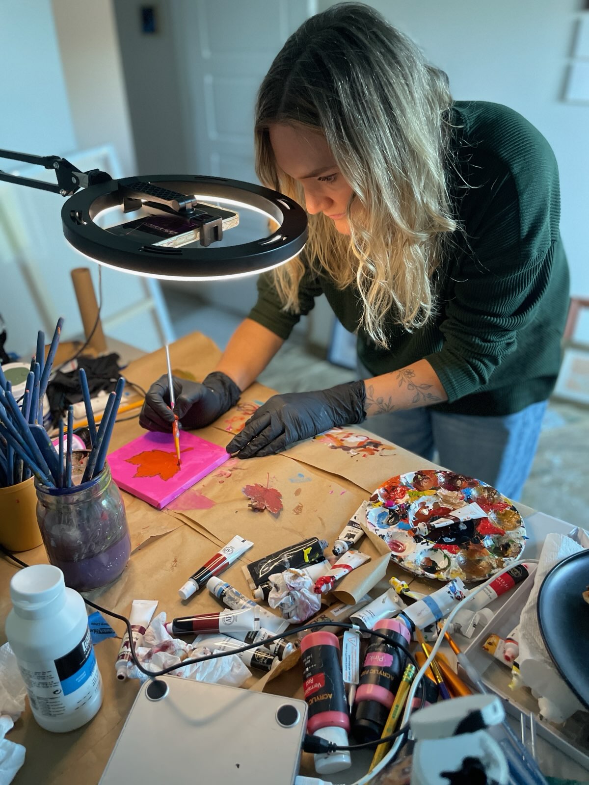 Rabekah painting a maple leaf at her studio desk, surrounded by brushes and paint
